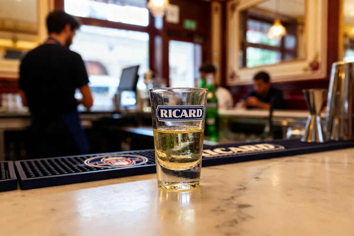 Verre de dose Ricard posé sur un comptoir de bar animé en arrière-plan
